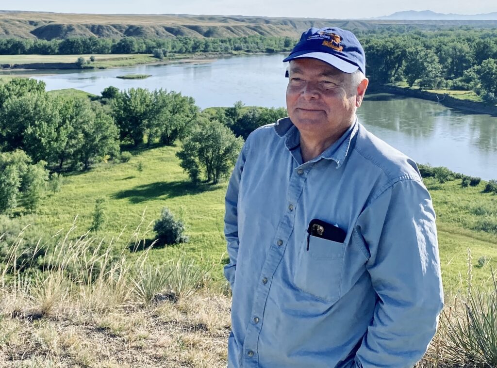 Smiling man in a blue cap and light blue shirt stands on a grassy hillside overlooking a river, trees, and distant hills.