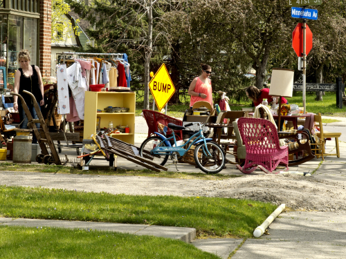 Outdoor yard sale on a sunny street with a clothing rack, yellow shelf, bicycles, chairs, and people browsing nearby.