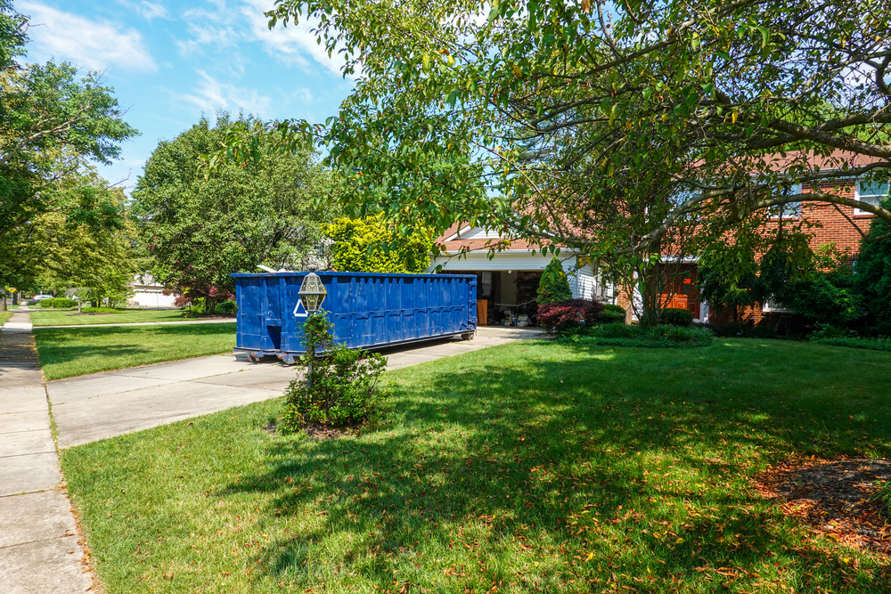 Blue dumpster parked on a concrete driveway beside a green lawn and a brick house.