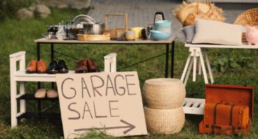Outdoor garage sale setup on grass with a table of cookware, shoes on a rack, baskets, a vintage suitcase, and a 'GARAGE SALE' sign with an arrow.