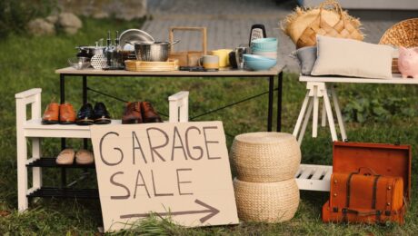 SignGarageSaleWrittenOnCardboardNearTablesWithDifferent Outdoor garage sale setup on grass with a table of cookware, shoes on a rack, baskets, a vintage suitcase, and a 'GARAGE SALE' sign with an arrow.