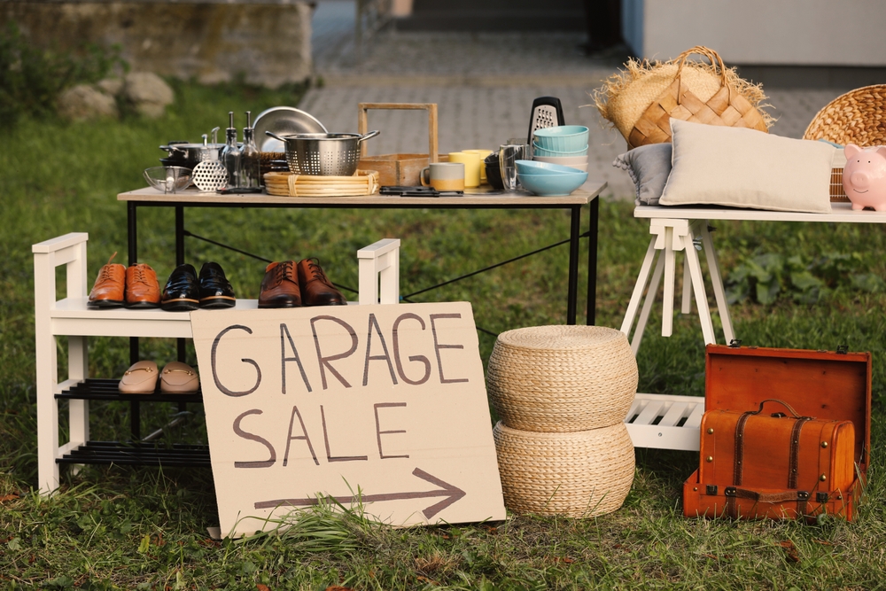 Outdoor garage sale setup on grass with a table of cookware, shoes on a rack, baskets, a vintage suitcase, and a 'GARAGE SALE' sign with an arrow.