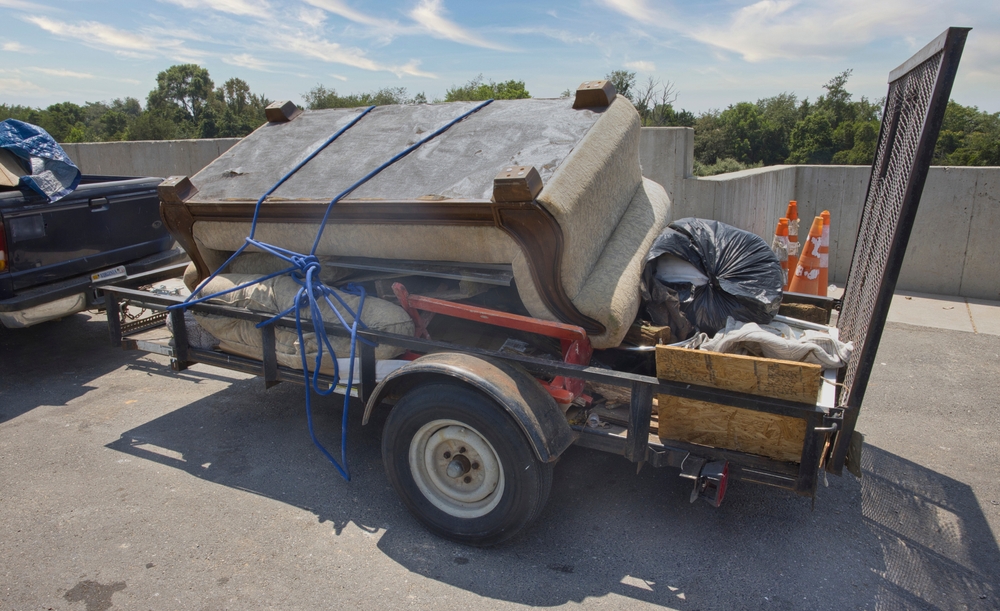 A trailer loaded with a beige sofa strapped down with blue ropes, plus a trash bag and wooden crates on the back.