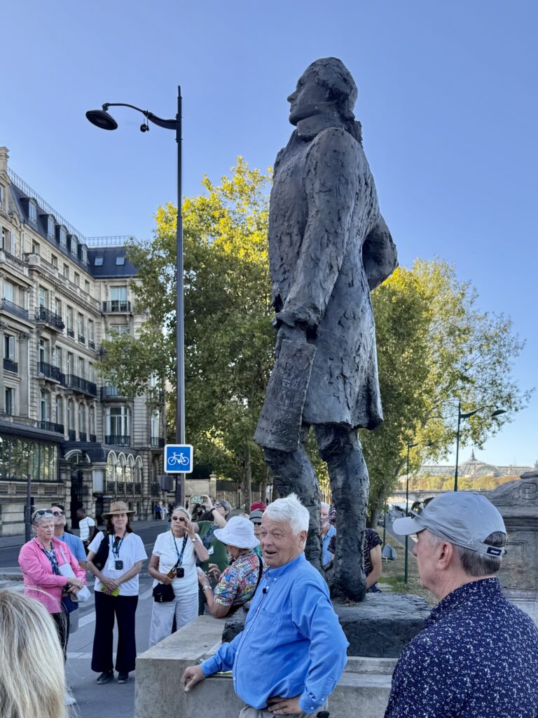 Life-sized statue of a man in a long coat in a city square, with pedestrians nearby and classic buildings in the background