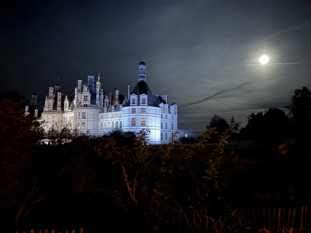 Castle-like mansion lit up at night with a bright moon in a cloudy sky, trees and a wooden fence in the foreground.