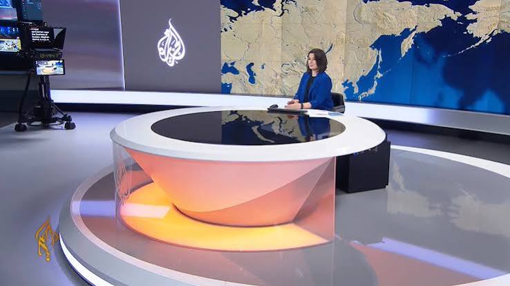 Female news anchor seated at a circular illuminated news desk in a studio with a large world map backdrop.